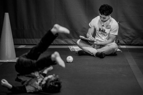 A grand valley student makes notes while a student from the childcare center lies on his back with his legs lifted in the air.