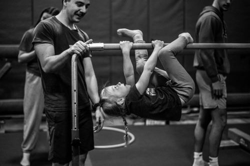 A small child shuffles along on an exercise bar with hands and legs wrapped around it. A grand valley student looks on.