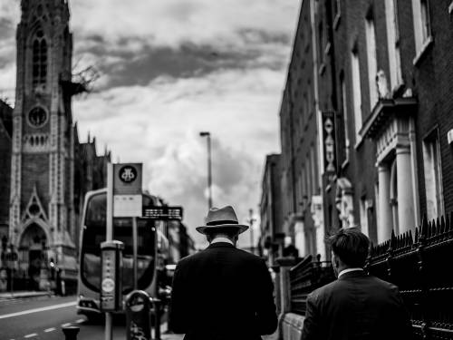 A black and white photo of the back of a man wearing a hat walking down a street in Dublin