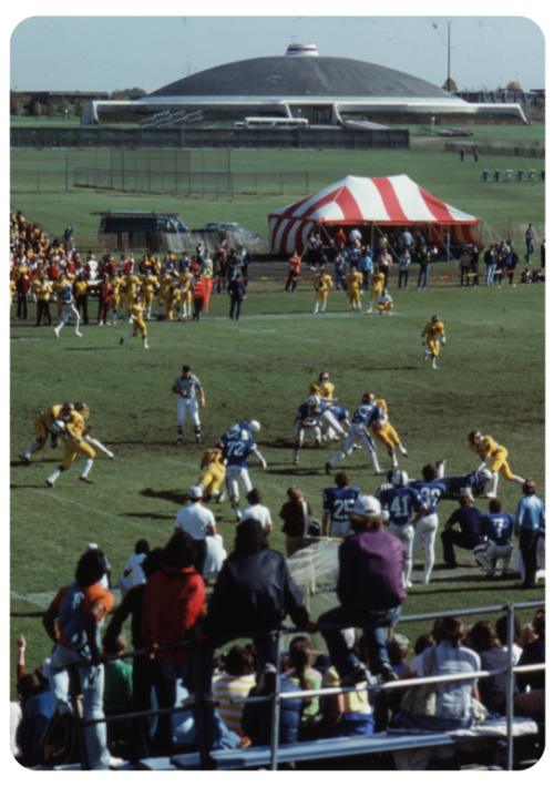 A football game in the 1970s, yellow and blue uniforms on the field. People sit on open bleachers in the foreground, in the background is a red striped tent and the dome.