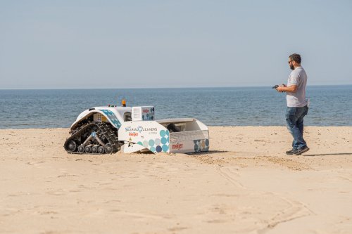 A man stands on a beach with water in the background. He is holding a remote control and is controlling a machine with the words "Searial Cleaners' and 'Meijer' written on it. 