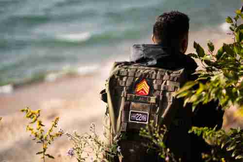 Camera is positioned behind Travis as he walks along the Lake. His camo backpack with patches from the army and mission 22 are prominent. 