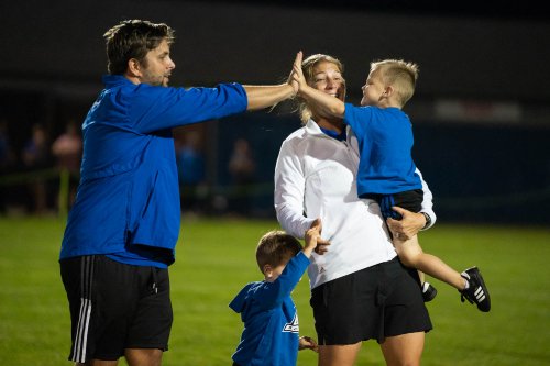 Coach Hultin holds one small child in her arms, and another by the hand. The child in her arms reaches out to high five Hultin's husband.