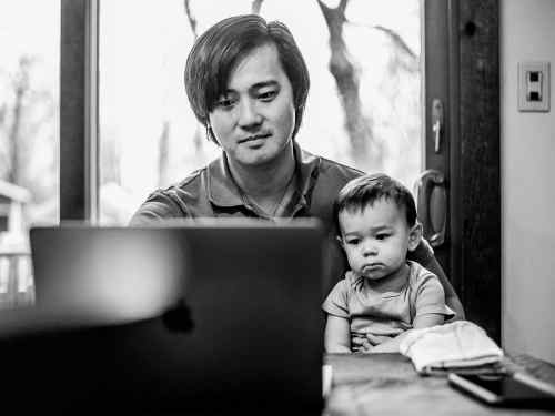 John Wen sits looking at his laptop while holding his young child on his lap