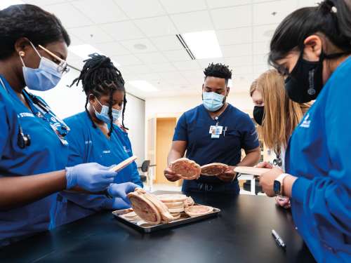 A group of students in medical scrubs and face masks stand around a table 