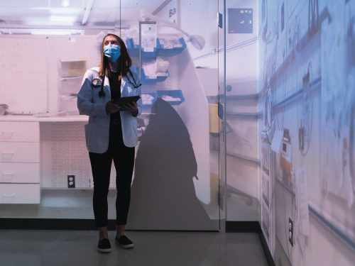 Woman stands wearing a medical coat, stethoscope and a face mask in a room with an image projected on the walls around her
