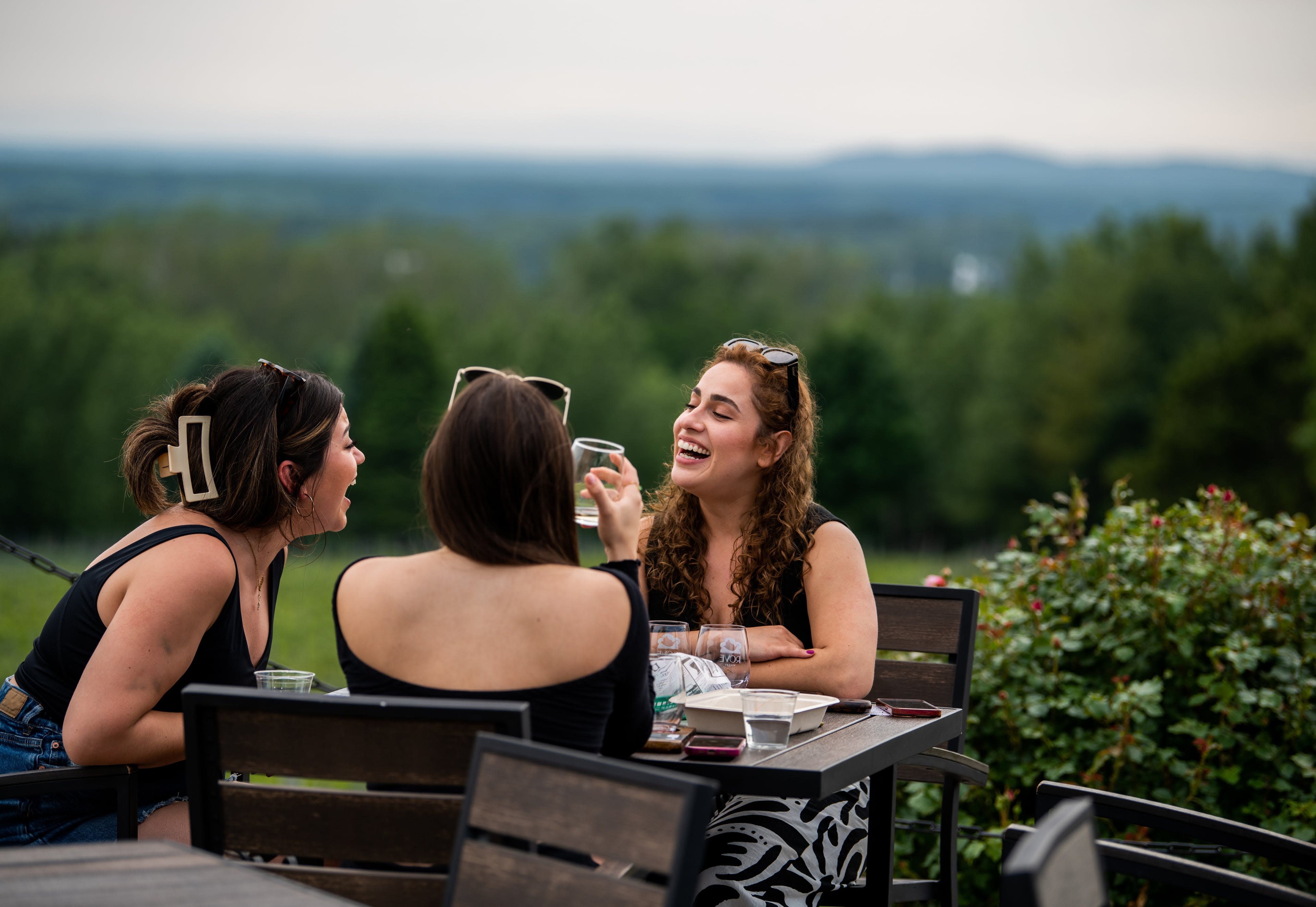 Three women sit outside on a patio while socializing, laughing and drinking wine