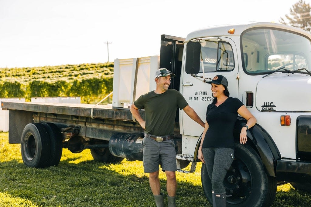 A man and a woman casually stand by a work truck in a vineyard