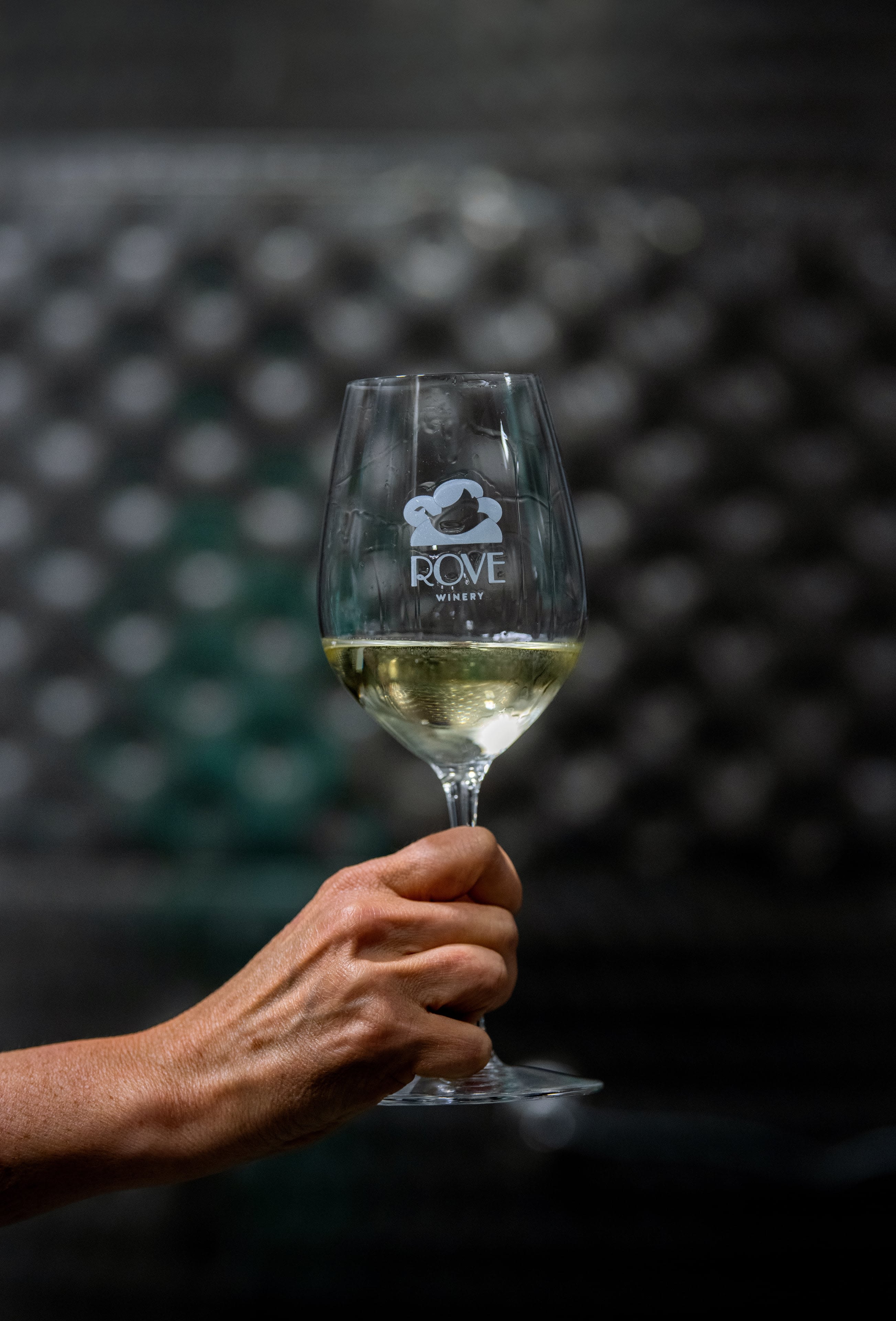 A close up photo a hand holding white wine and a Rove winery logo etched on the glass
