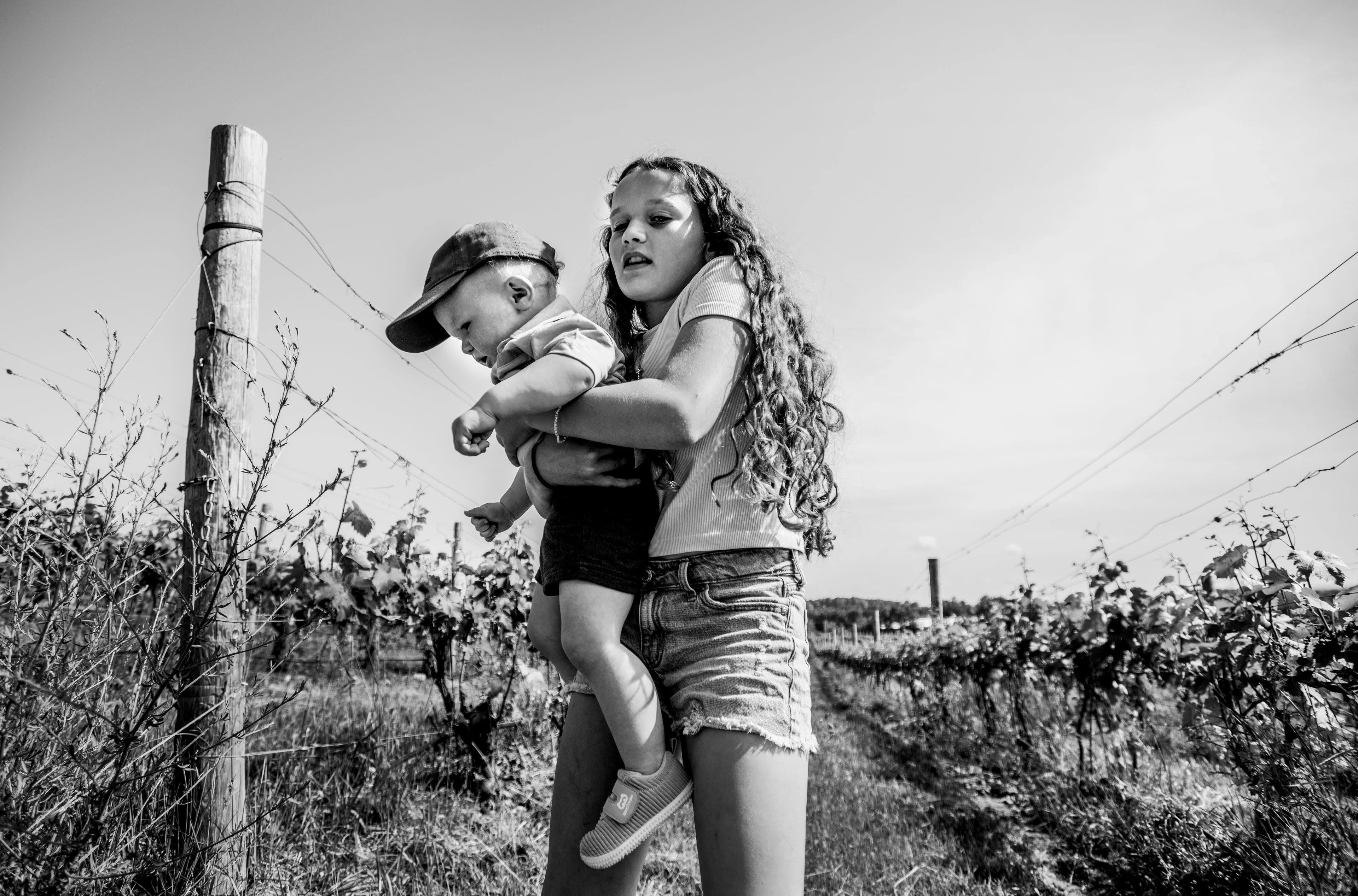 A black and white photograph of a young girl holding a toddler in her arms while walking through a vineyard