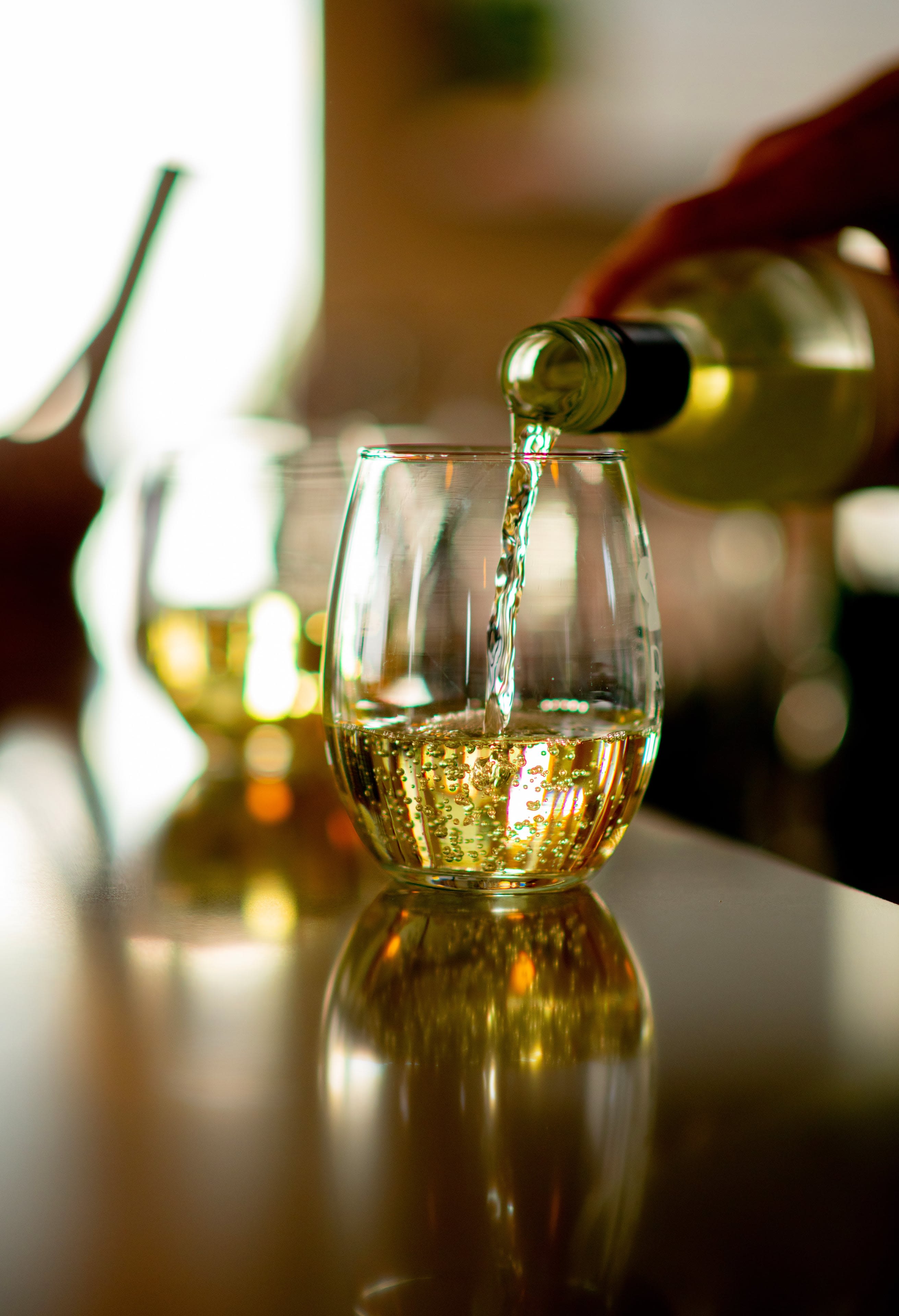 A close up photo of a stemless wine glass being filled with white wine, the light behind the glass makes the wine glow a lightgold