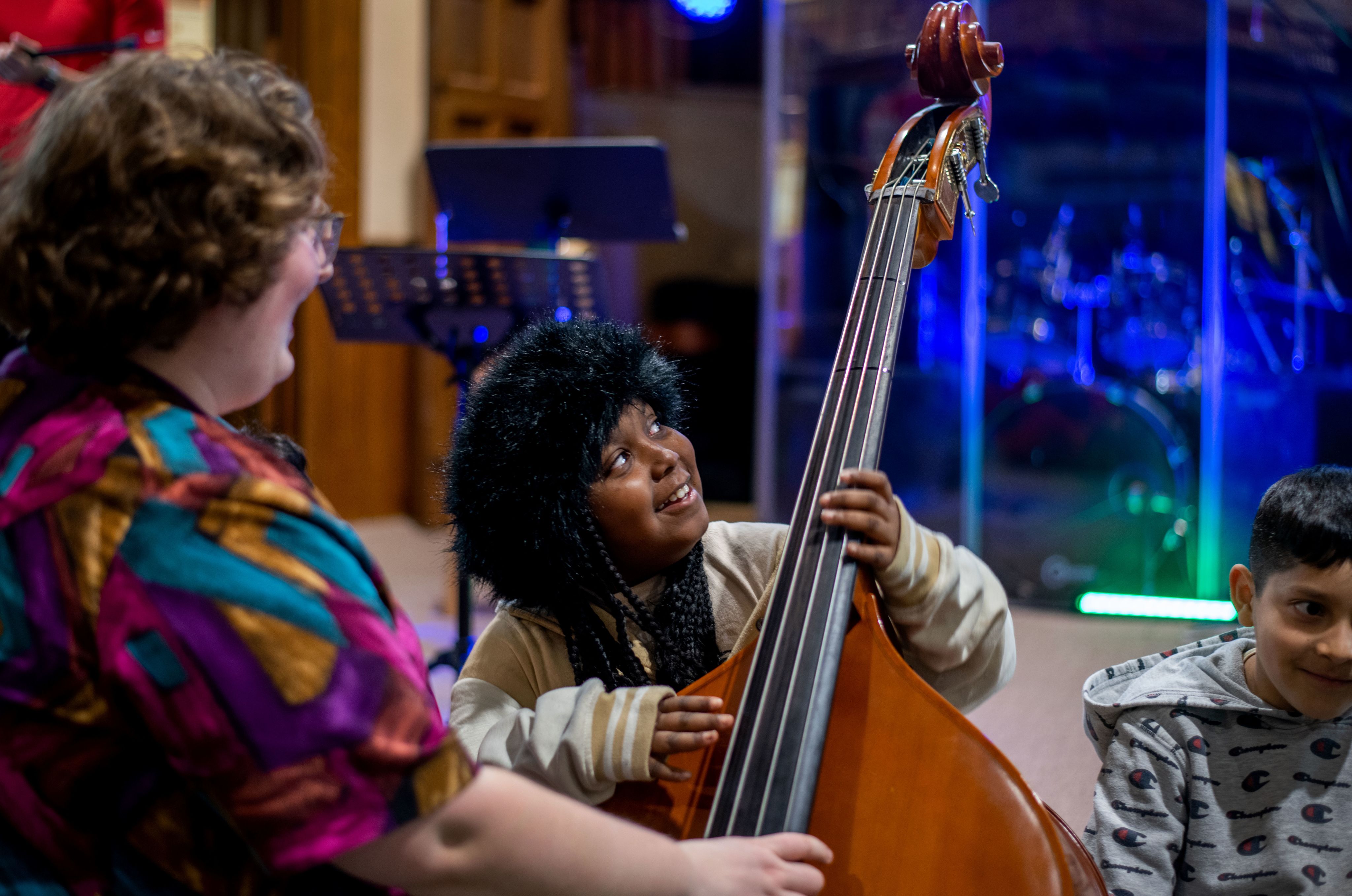 a young student holds a double bass smiling up at the instrument, a woman in print shirt smiles and looks on