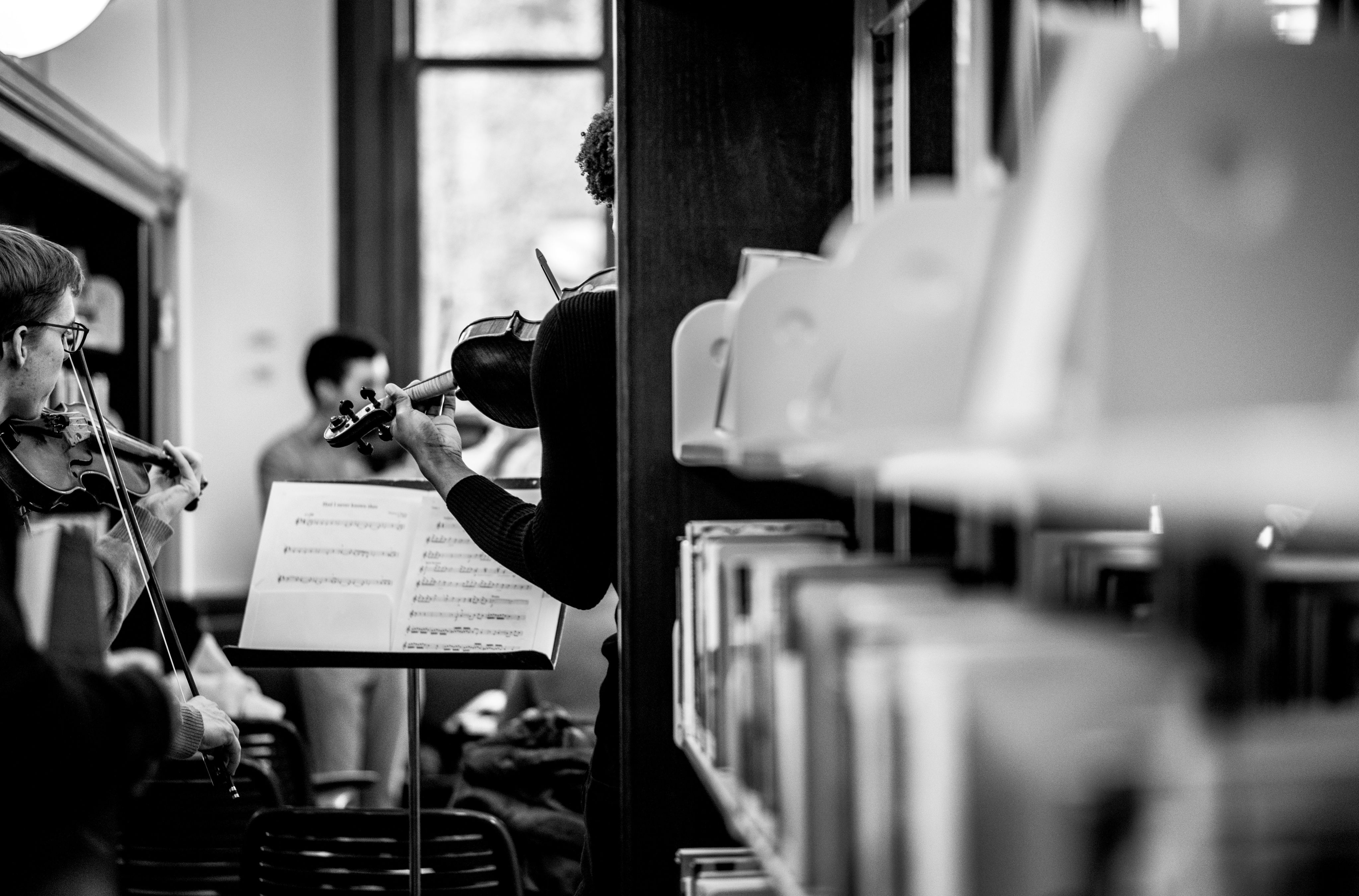 black and white of string musicians playing at a library, book stacks visible in foreground