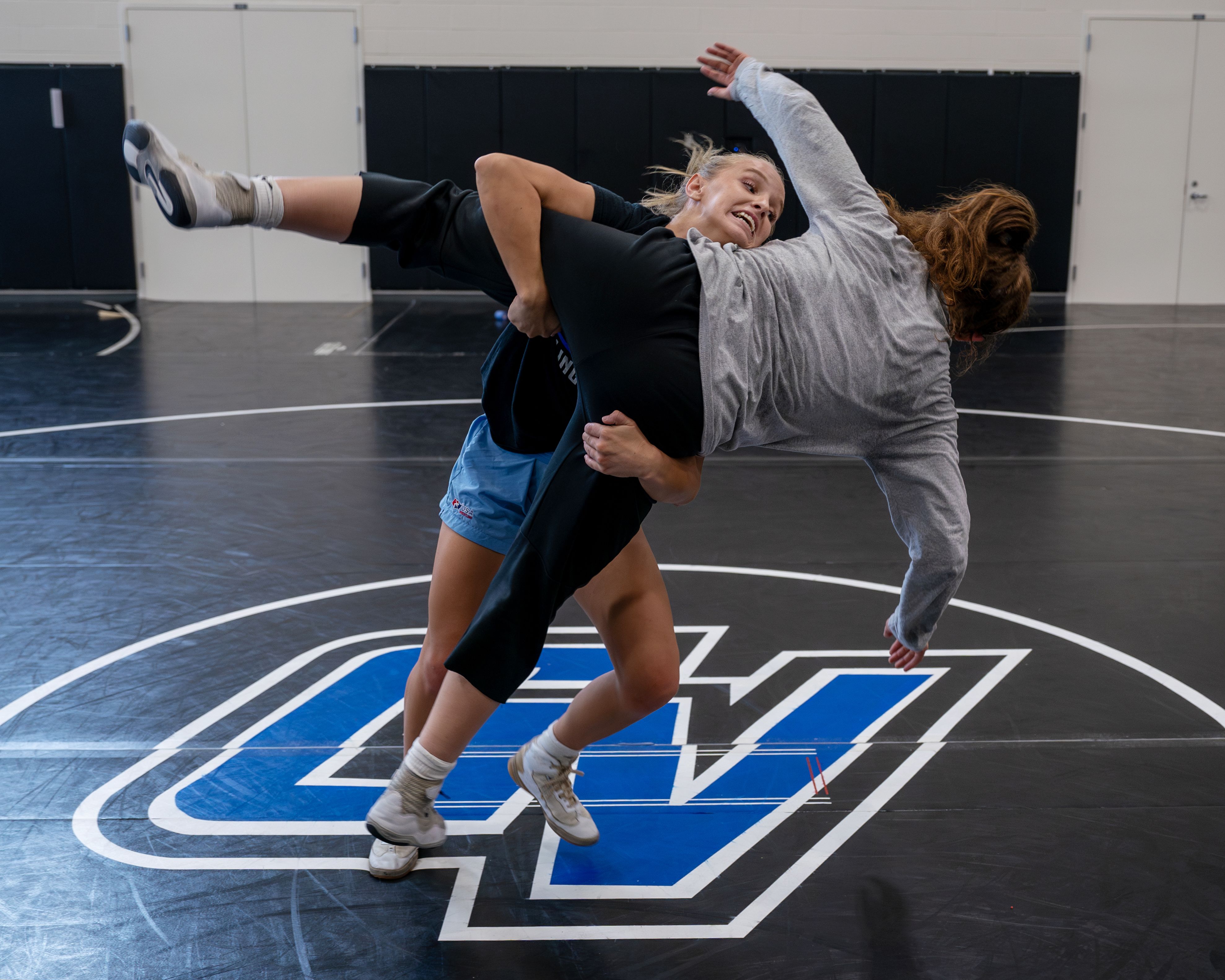 one woman wrestler takes another to the mat, which has a GV in the center