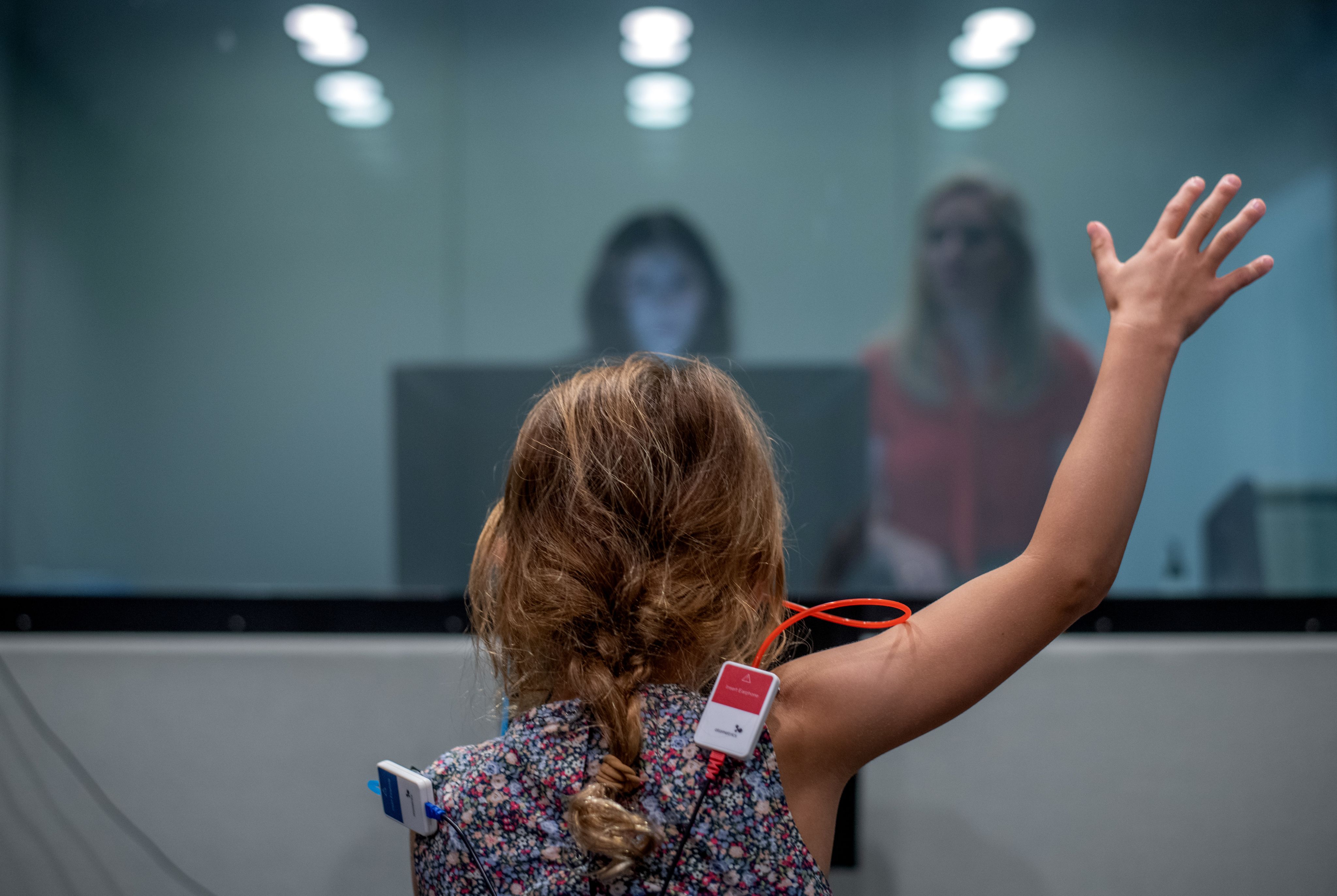 young girl raises hand during hearing tests in room, while two people stand in another room looking at her while conducting testing