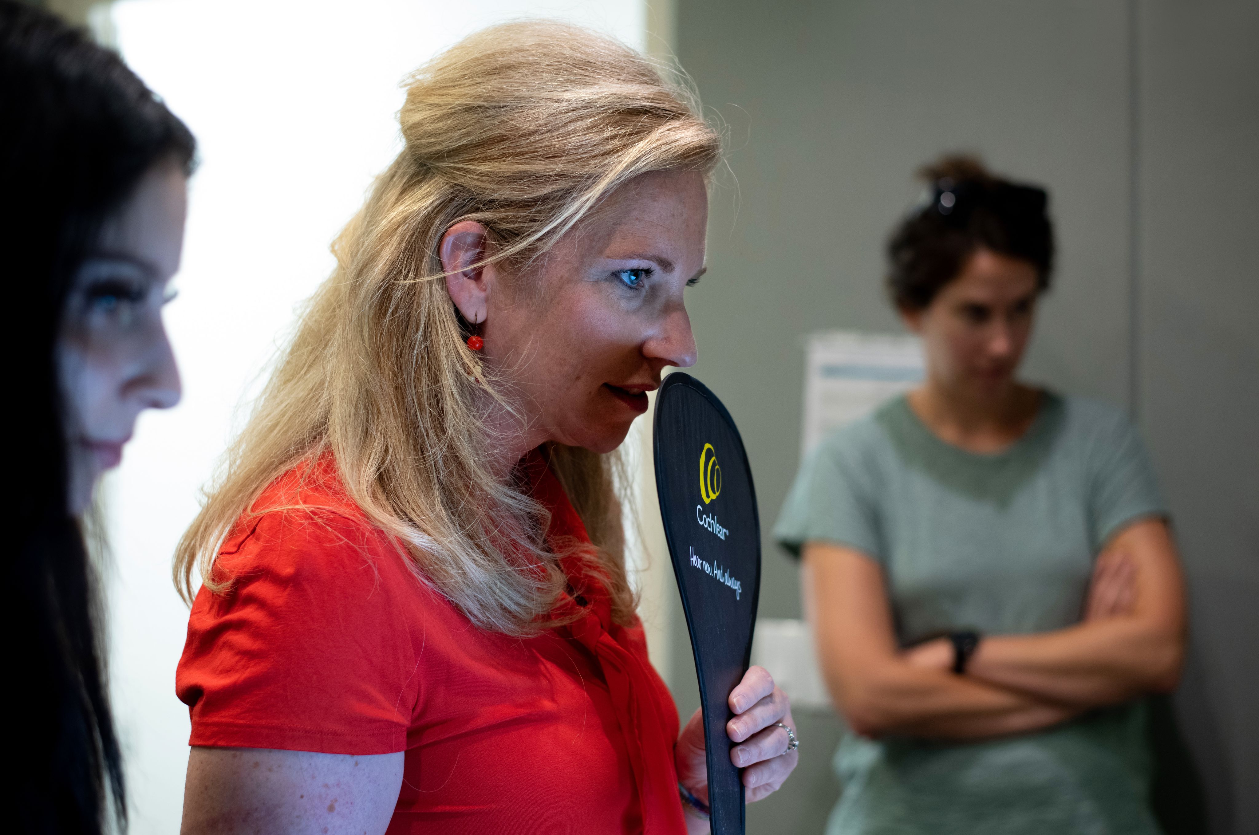 Three women look off camera, the one in the middle in red holds up a black paddle.