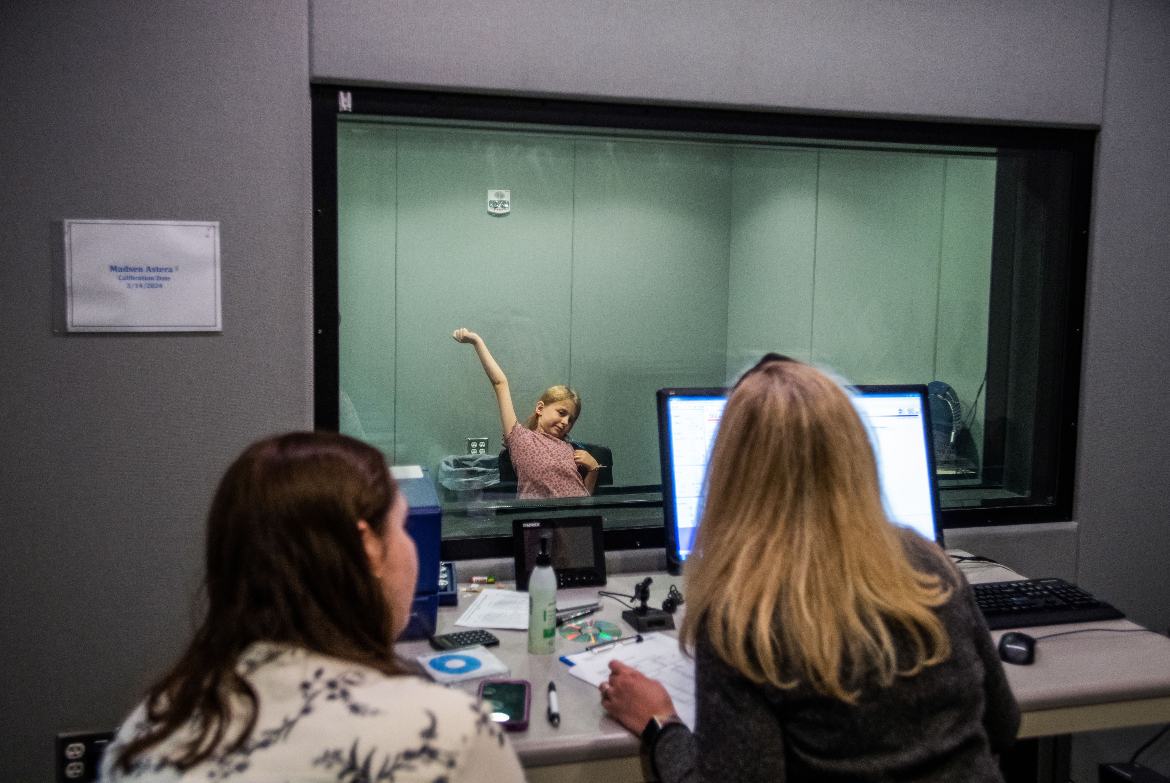 girl raises hand while seated in a hearing testing room, two people look on over computer monitors in another room