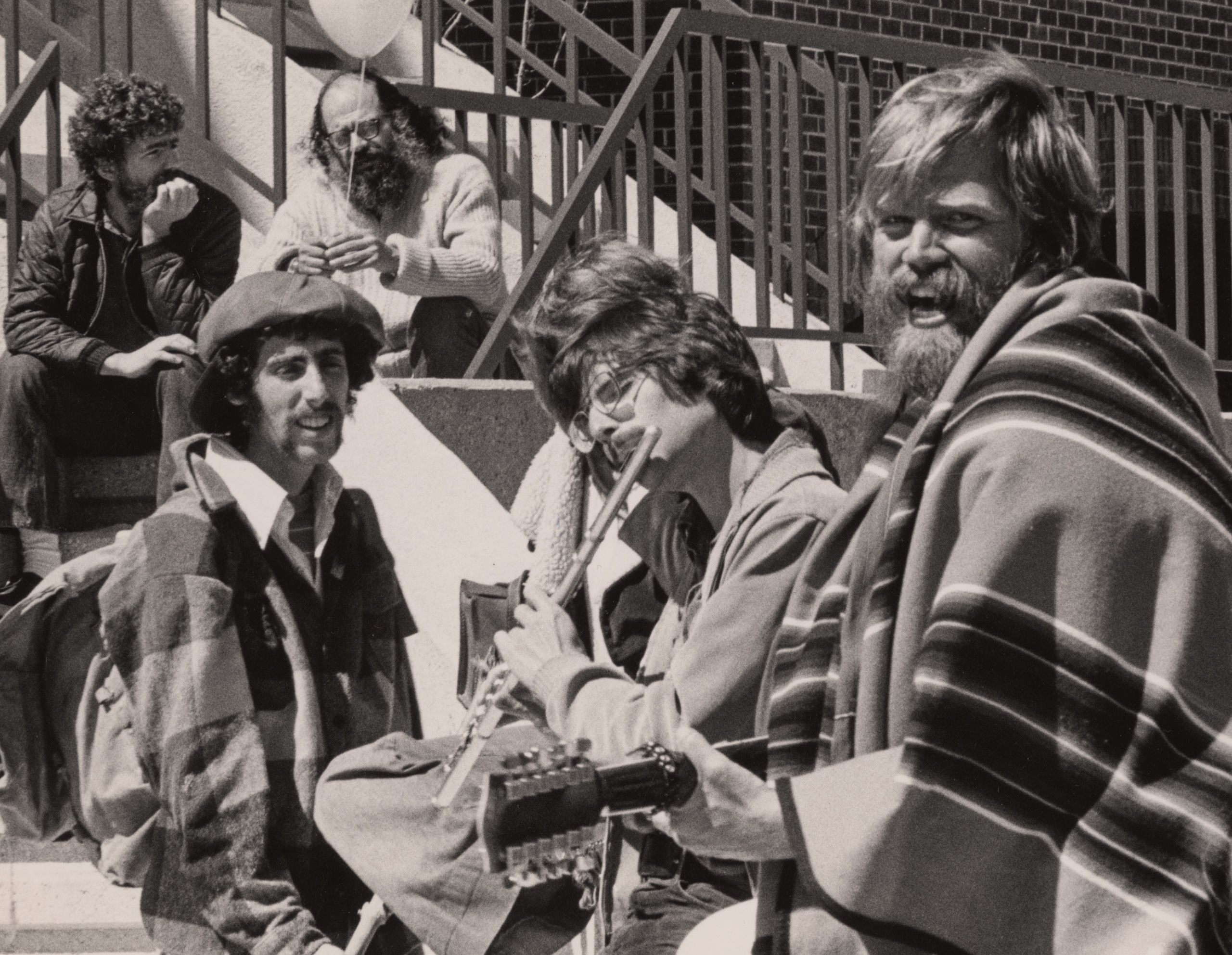 black and white photo of 1970s GVSC, with people seated outside, one man plays the flute