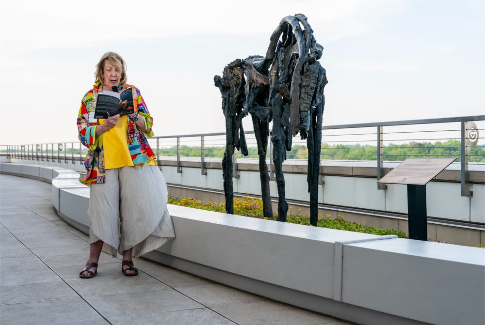 woman reading from book with microphone next to horse sculpture created from wood