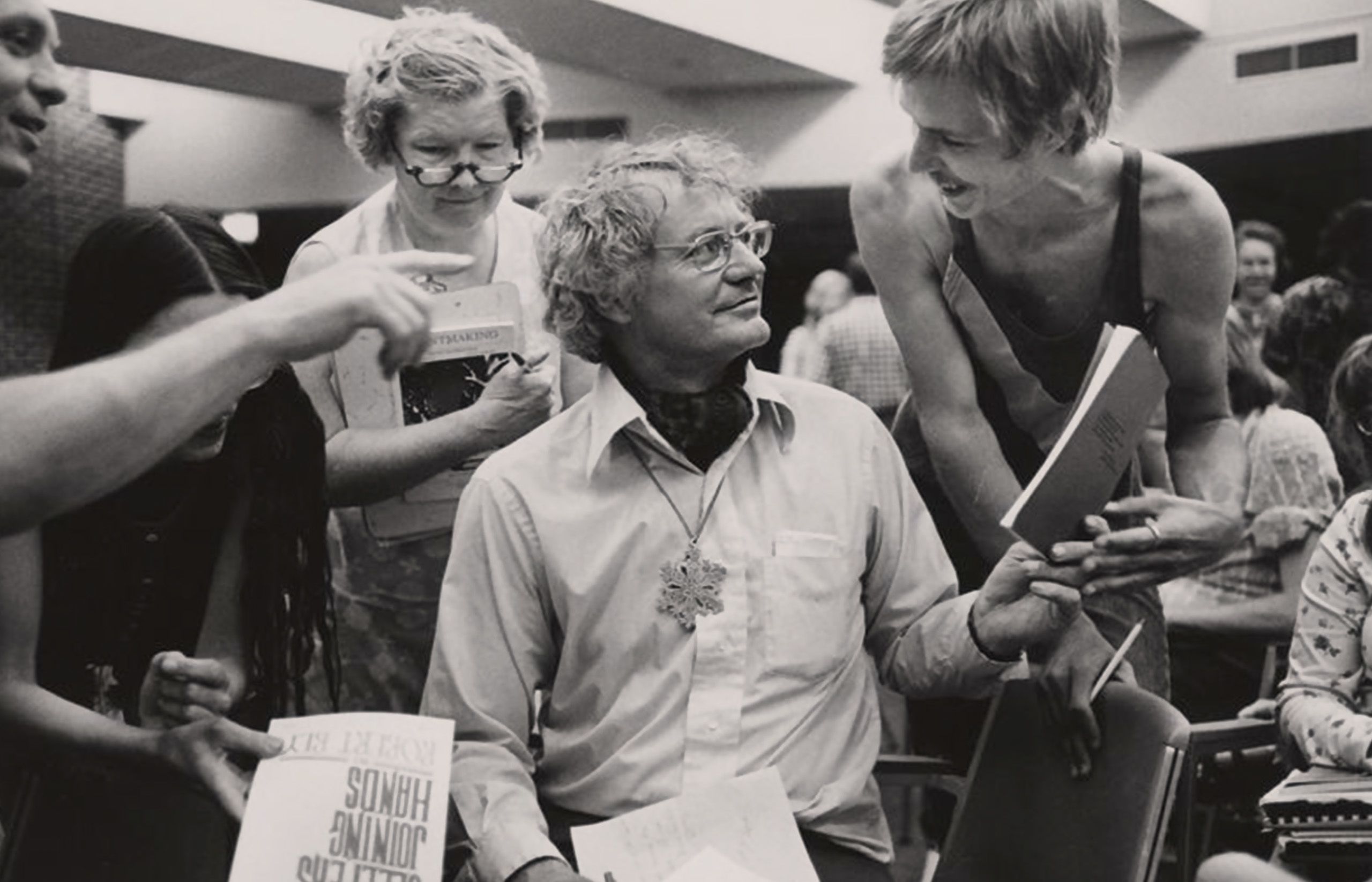Robert Bly in center, women crowded around him with papers and books, black and white photo