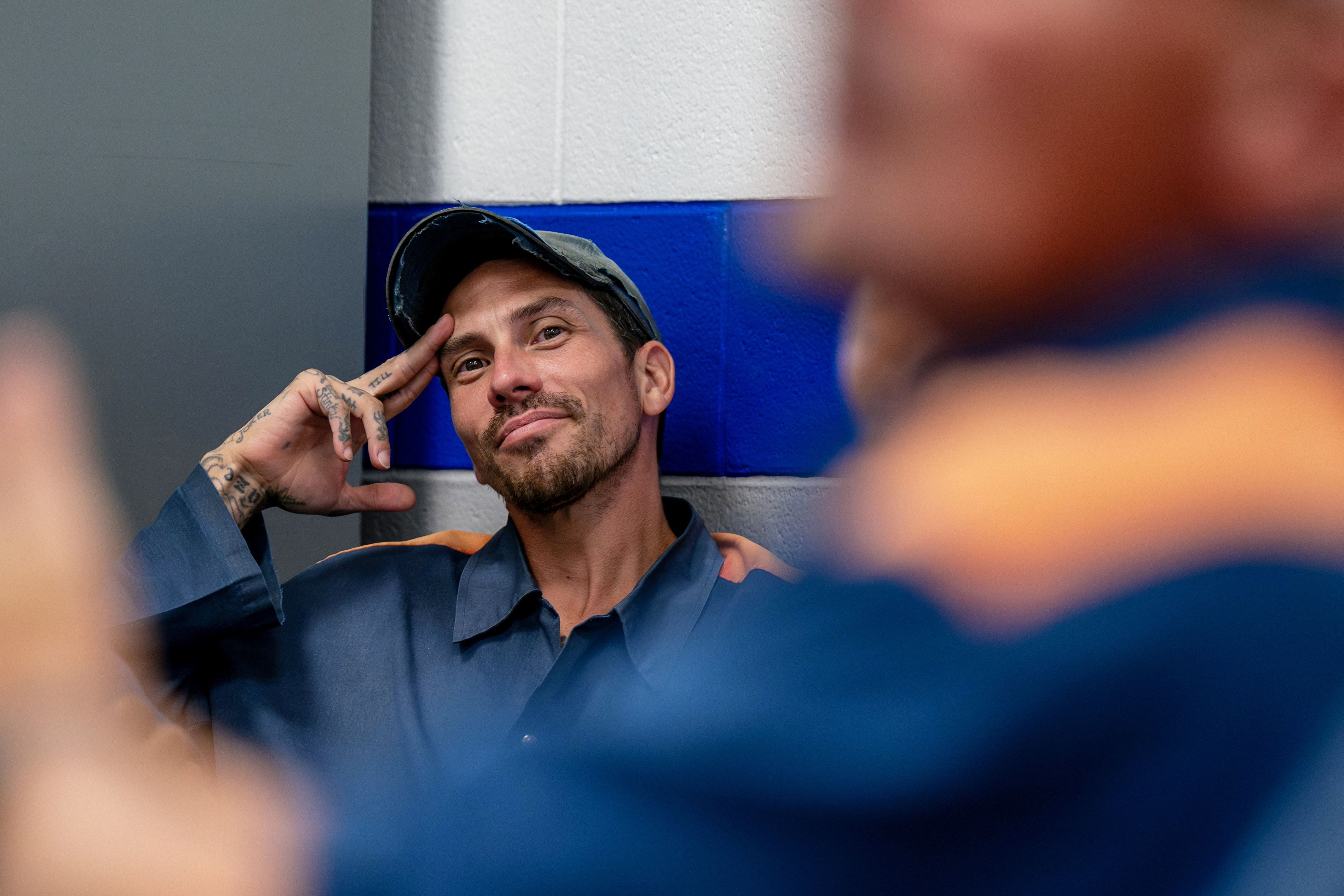 inmate in blue and orange uniform in back of class looks thoughtful with head resting on hand and wearing ball cap
