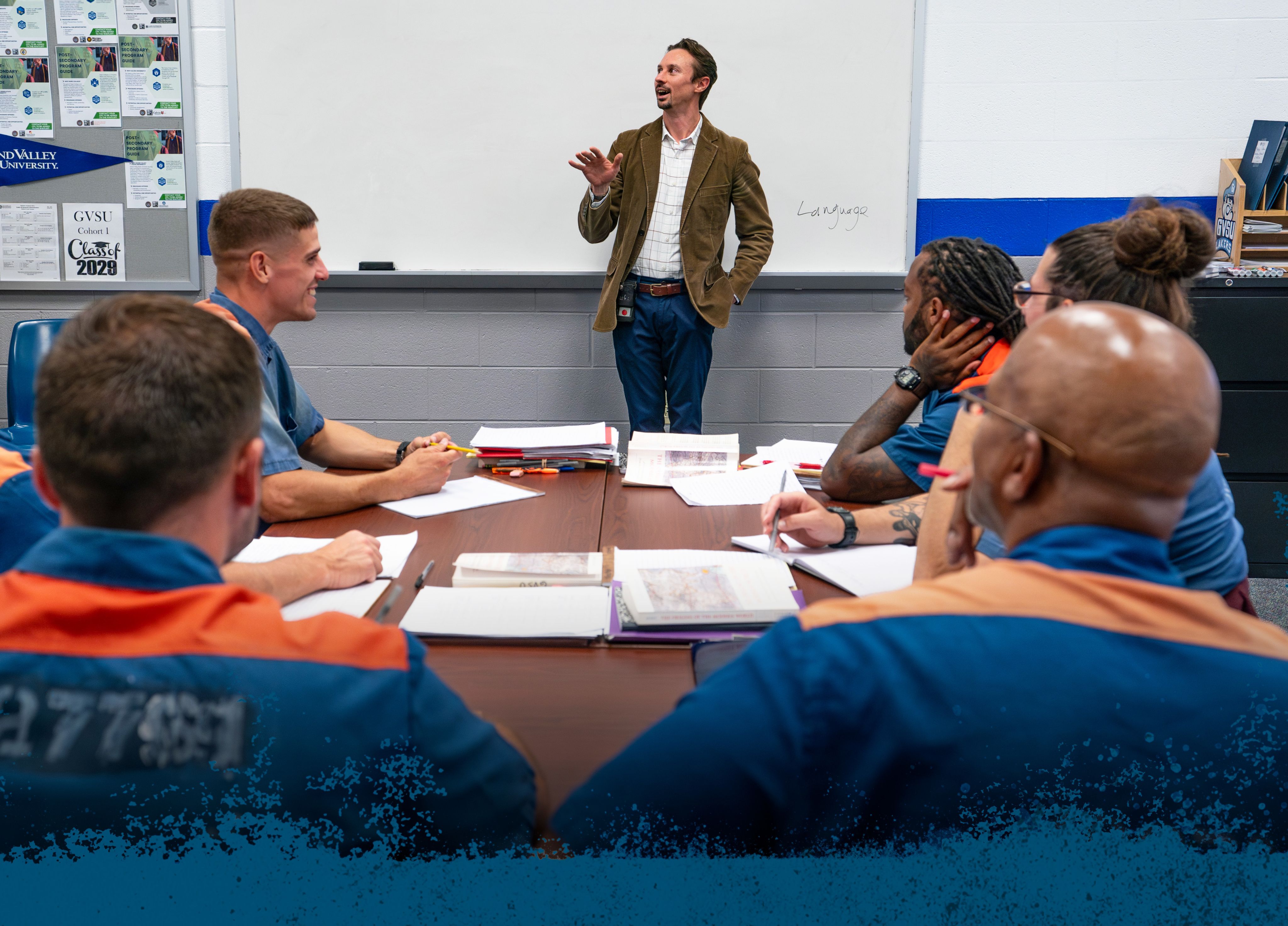 Aaron Yore-VanOosterhout stands in front of whiteboard in classroom, table of six inmates in blue and orange uniforms smiling, interacting
