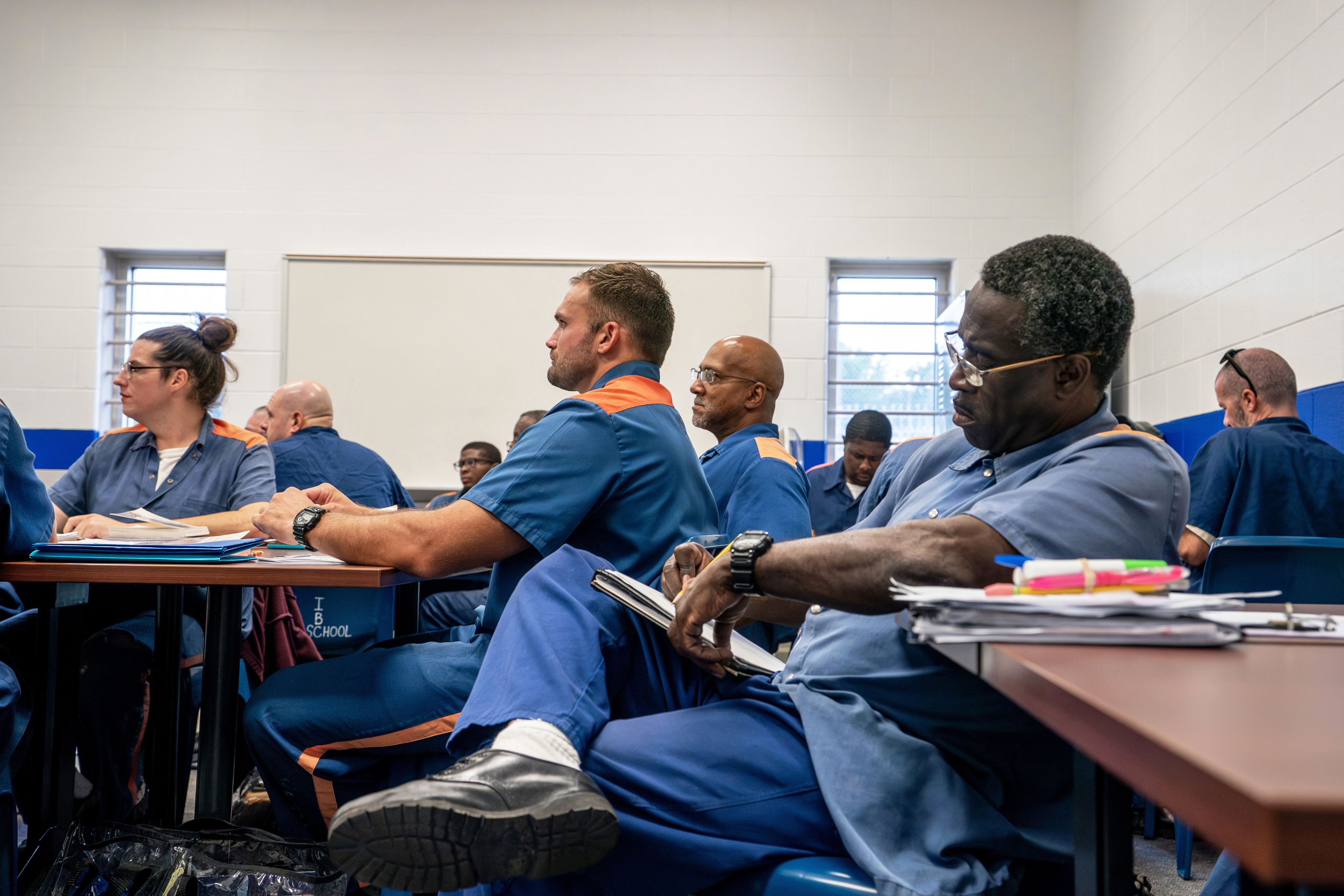 classroom of tables, inmates in blue and orange uniforms work on papers or look up at front of room