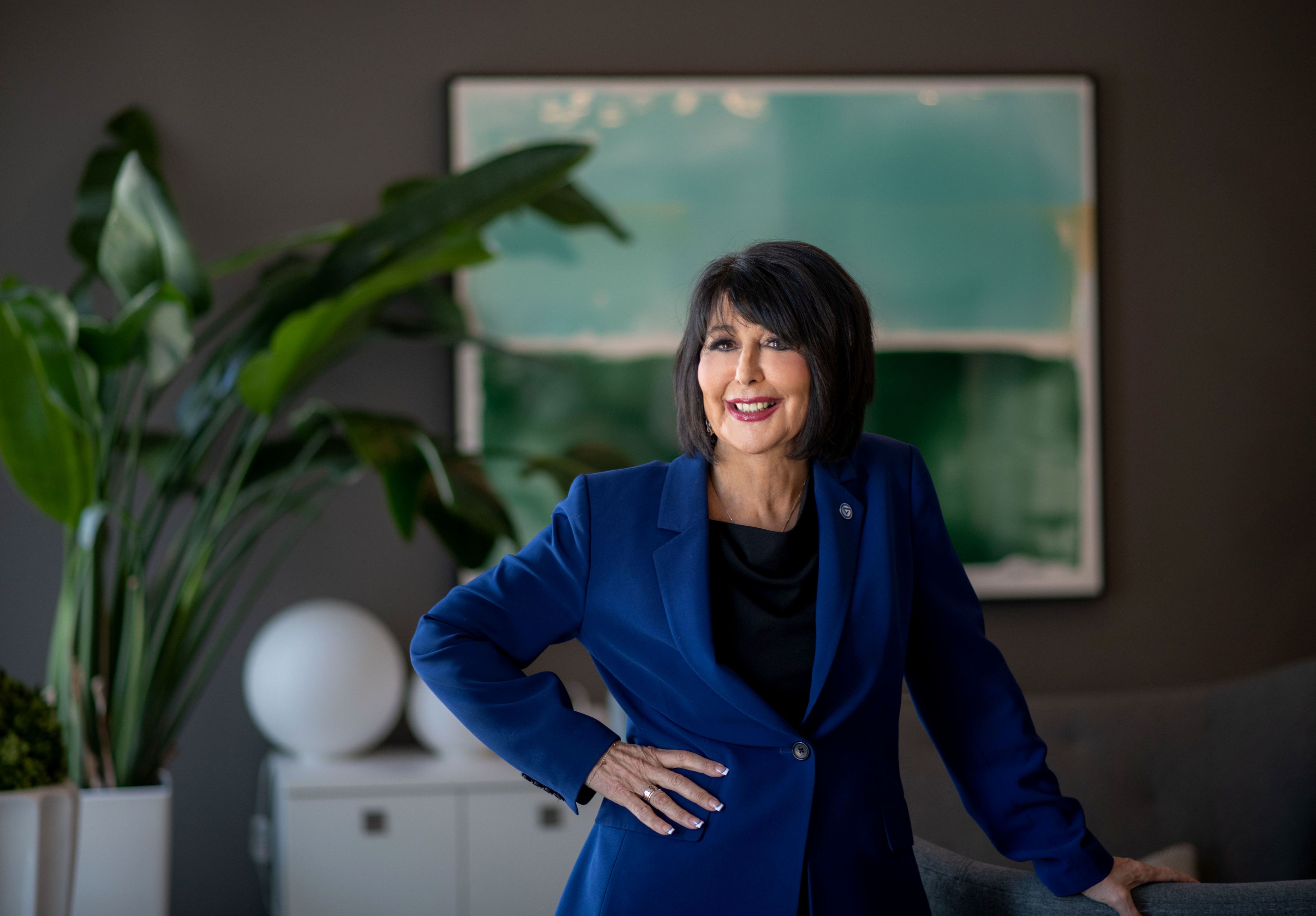 Philomena V. Mantella stands with a hand on hip and in a blue blazer. A green and blue painting and green plant fill in the background.