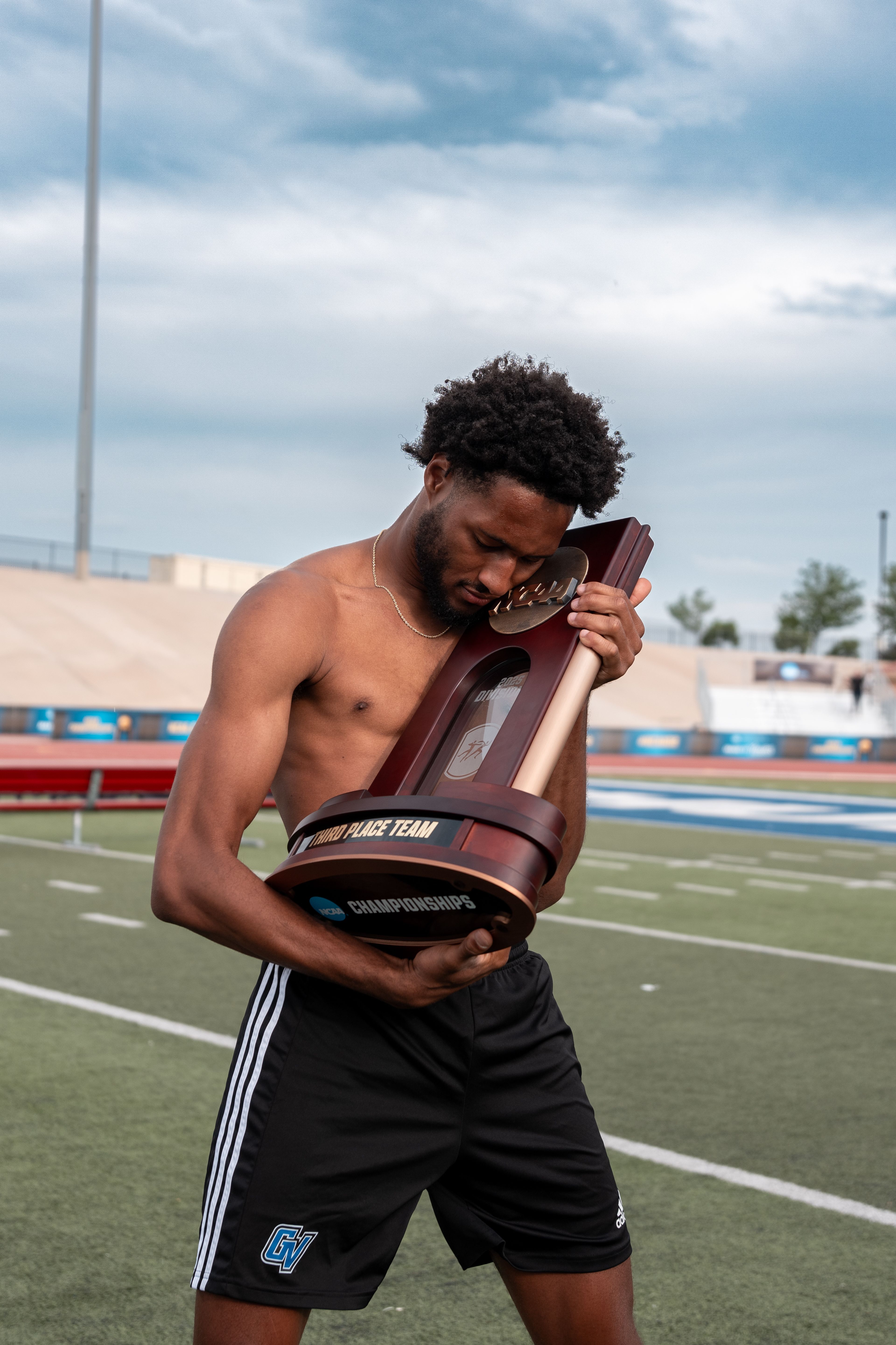 male track athlete without shirt on hugs a NCAA championship trophy