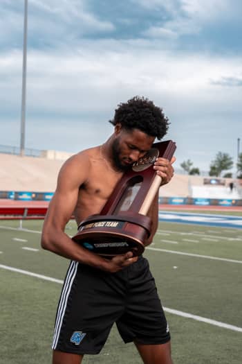 male track athlete without shirt on hugs a NCAA championship trophy