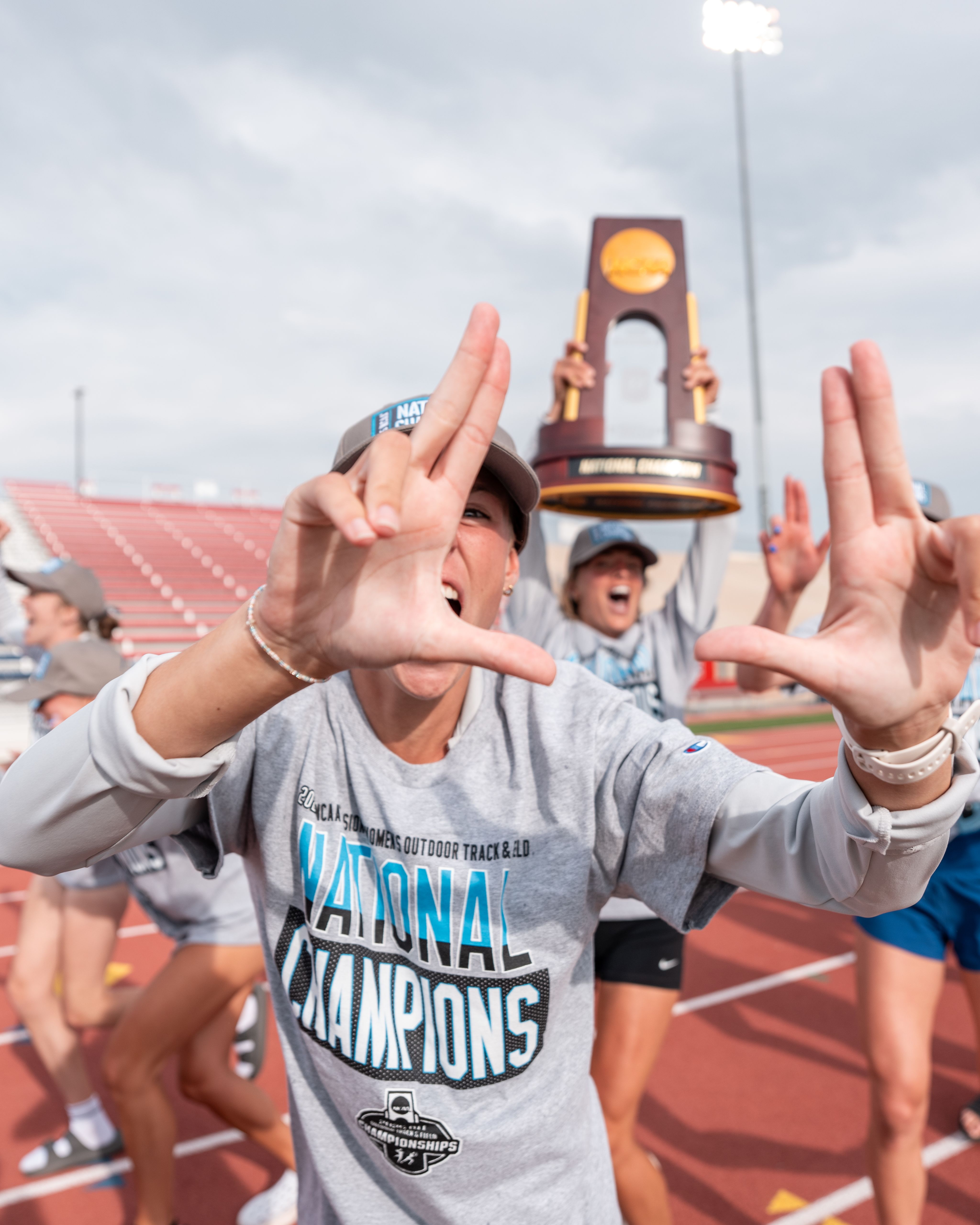 track athlete makes hand gestures after win, person in back holding NCAA trophy high above head