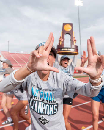 track athlete makes hand gestures after win, person in back holding NCAA trophy high above head