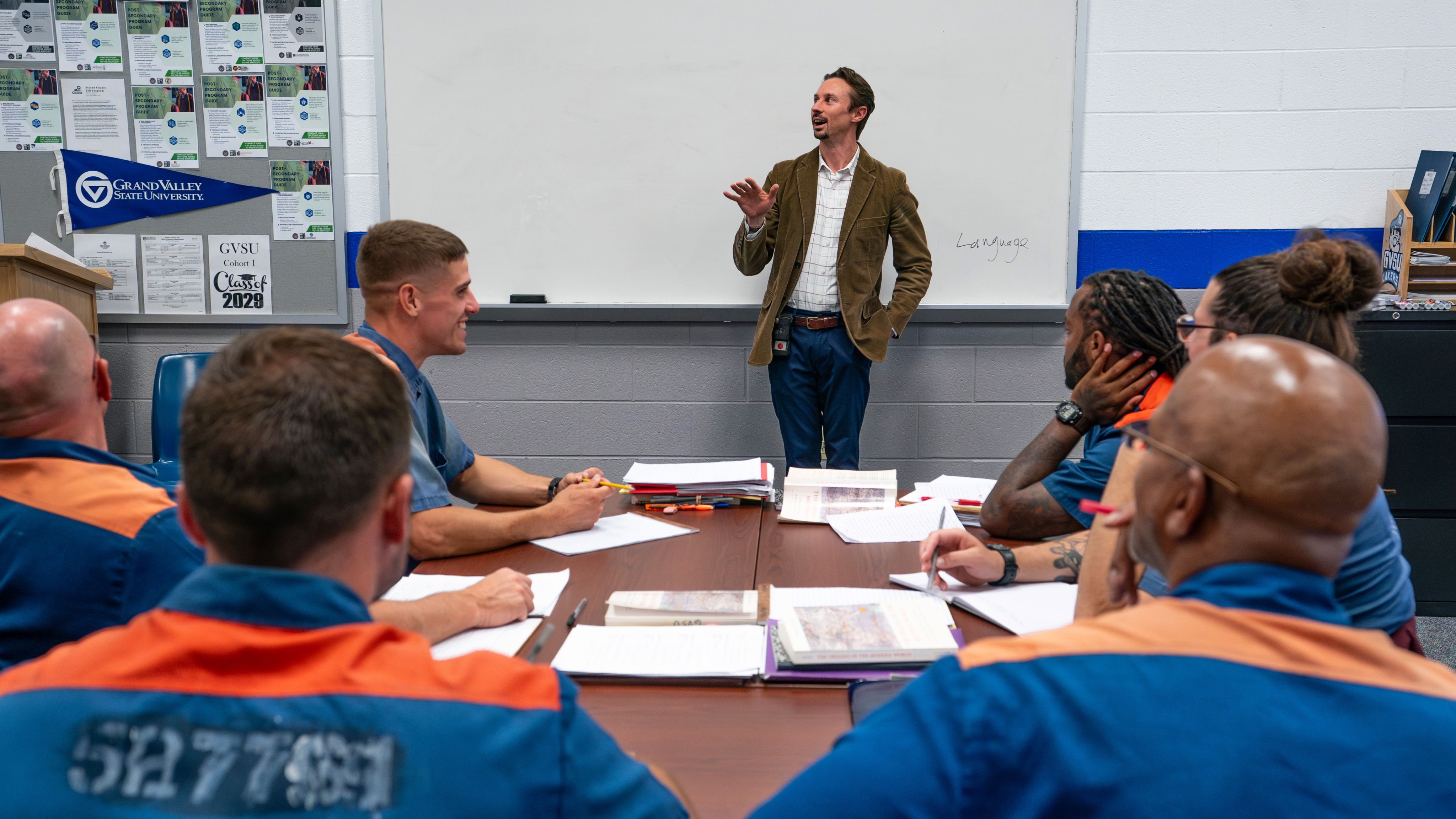 class of inmates in orange and blue uniforms, man in brown jacket at front of class near whiteboard