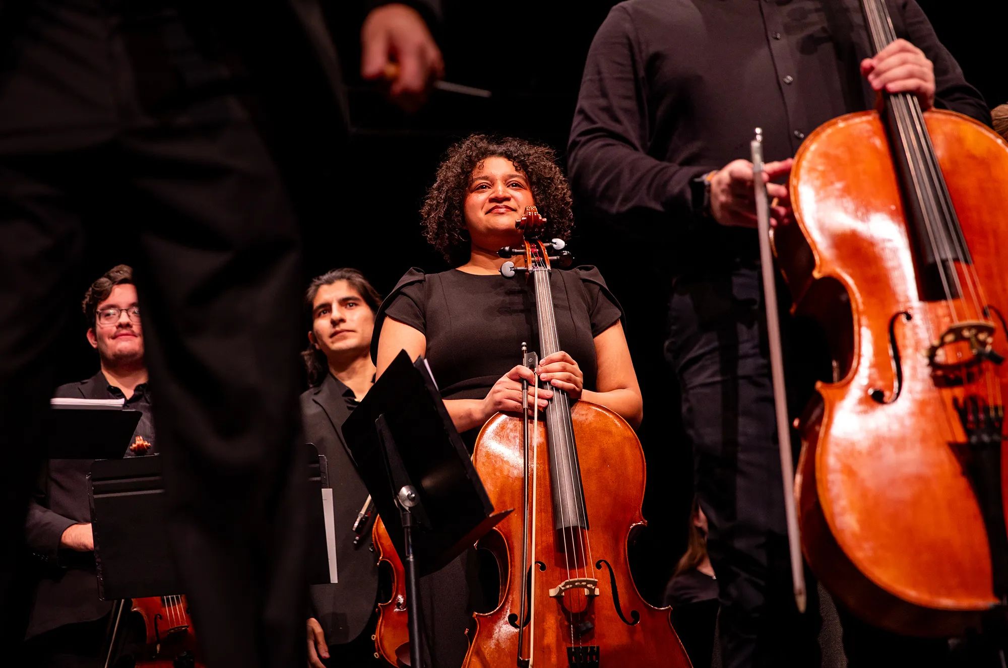 Maria Benítez-Sabino stands behind her cello, other musicians and music stands pictured; everyone dressed in black