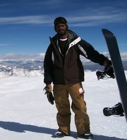 Thomas Pikes stands outside wearing snowpants and a ski jacket, holding a snowboard