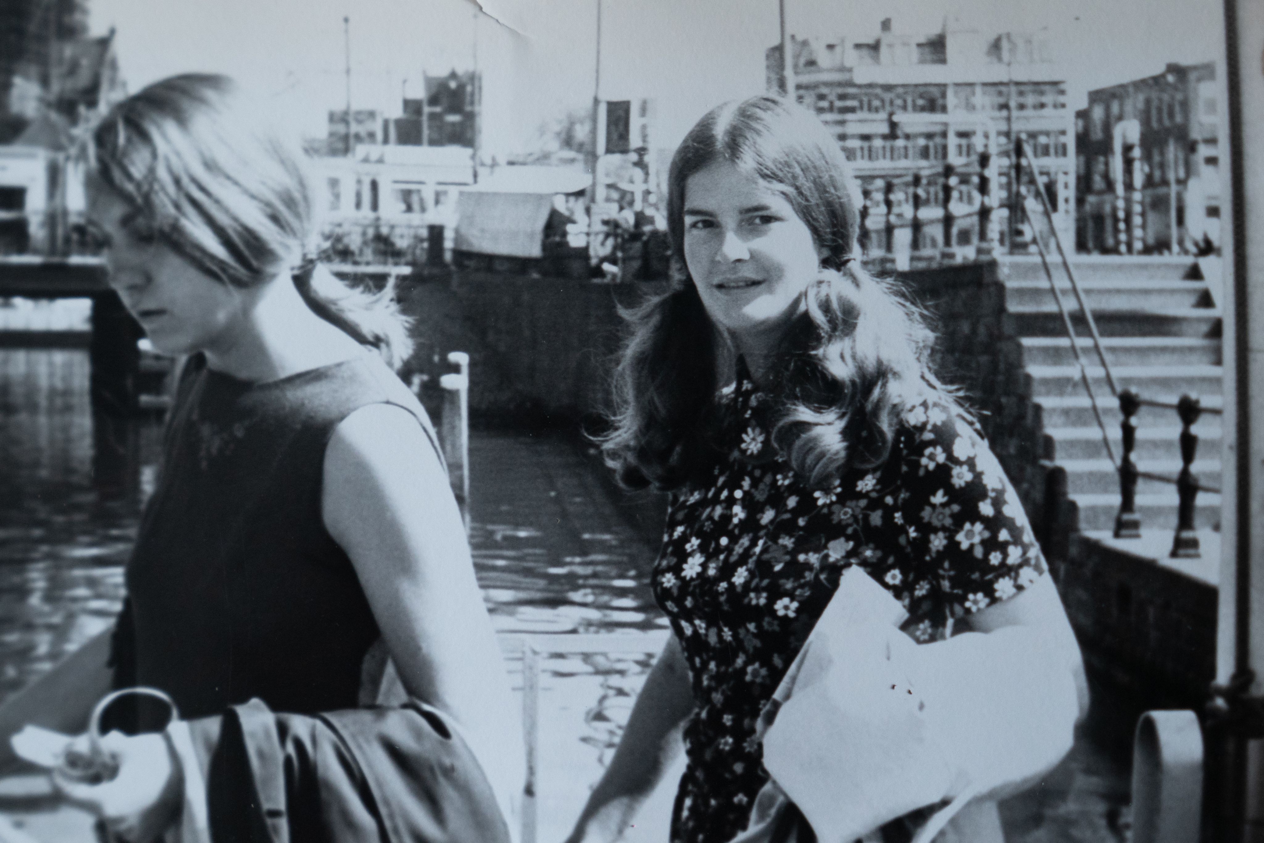 Two young women are standing with water in the background. Stairs to the right lead down to where they are standing. A city is in the background.