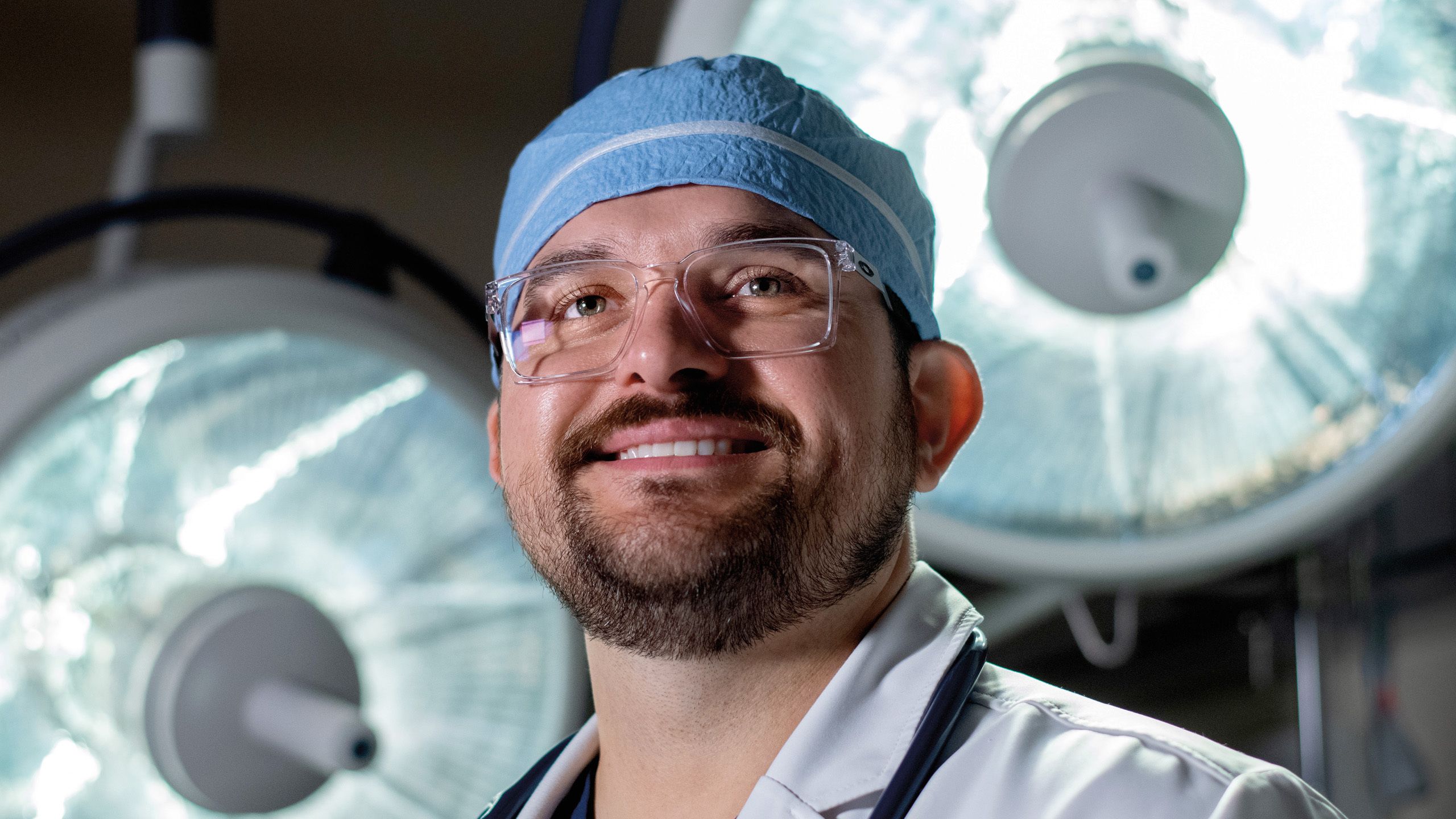 Man in a light blue medical cap and doctors coat wears clear glasses and smiles. Surgical lights are behind him.