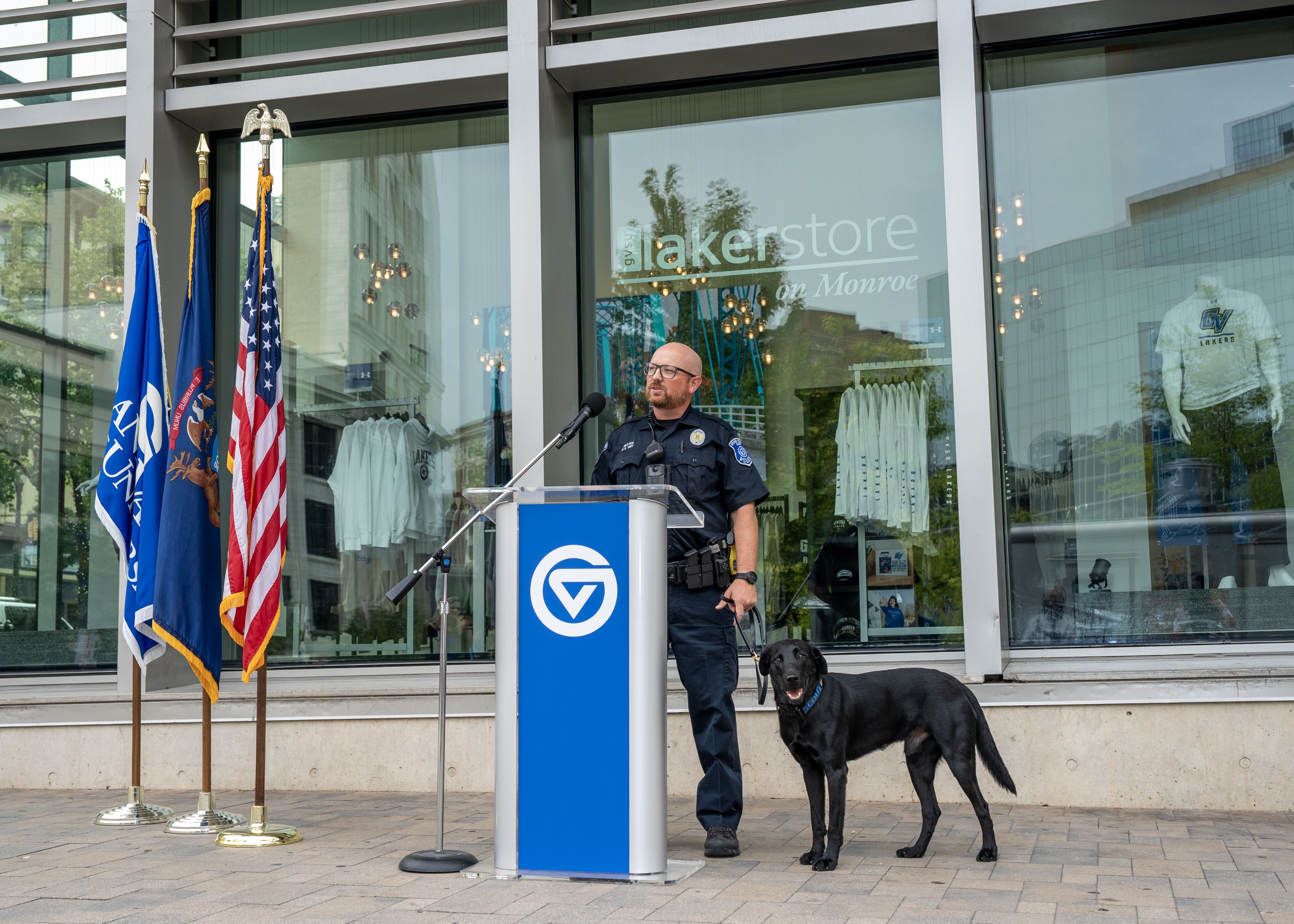 Officer Paul Weaver stands at podium with Scout on leash by his side, in front of GVSU Laker Store downtown