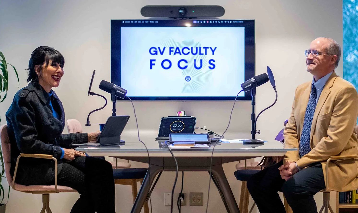 President Mantella, left, and Matthew Daley sit at a table with podcast microphones; GV Faculty Focus on the screen in back
