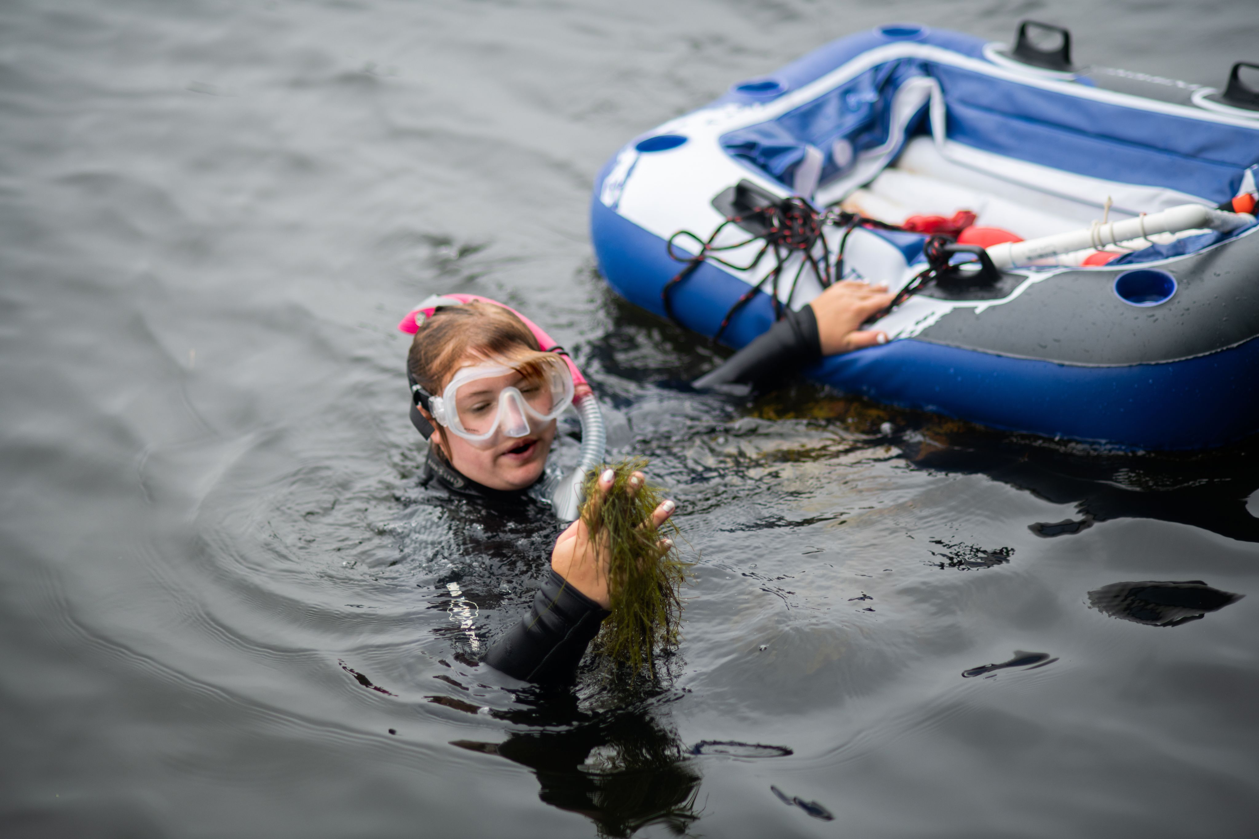 a student in a wetsuit holds algae in one hand while holding an inflatable raft with the other