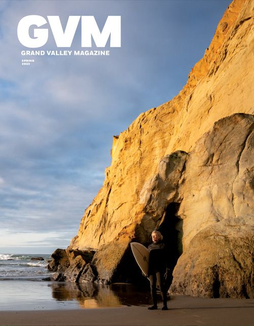 A man with a surfboard stands on the shores of a beach. In the background is an ocean and a big sand colored cliff. Text reading "GVM" is in the top left.