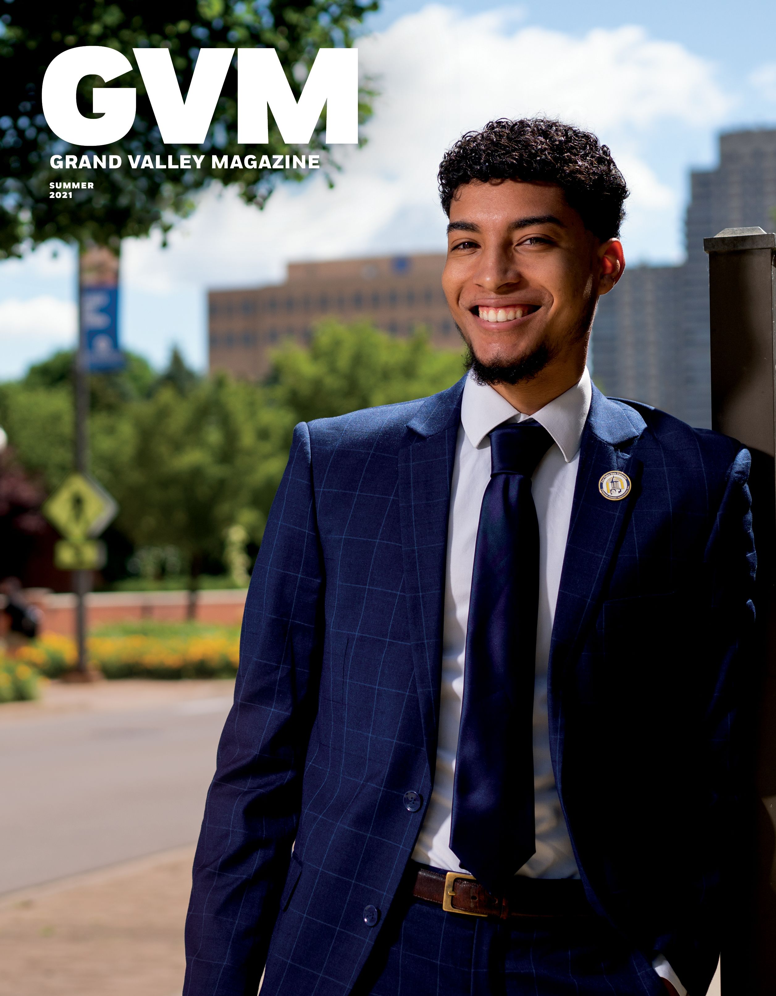a Black student wearing a navy blue suite standing a crossed the street from the Richard M. DeVos Center with the WGVU Building in the background