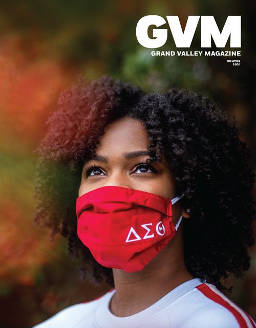 African American woman looks upward. She wears a red mask with her sorority emblems on it. The background is blurred fall trees.