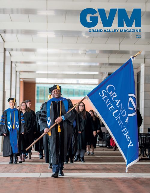 Faculty and staff in black and blue robes walk through the covered walkway connected to kindschi. Felix Ngassa leads the group with a black cap and yellow tassel and holds a blue flag with a gvsu logo.