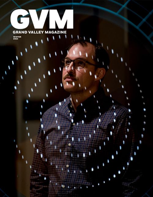 A man with a mustache and glasses looks off camera. He's wearing a plaid blue button up. Artistic lights are layed over top of the image in a swirl pattern made up of white dots. The letters GVM are in the top left corner.