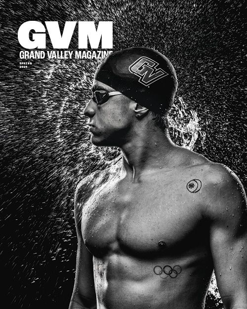 A black and white photo of a male swimmer wearing goggles and a swim cap with a Grand Valley logo on it looks left with water droplets spraying all around his head and torso