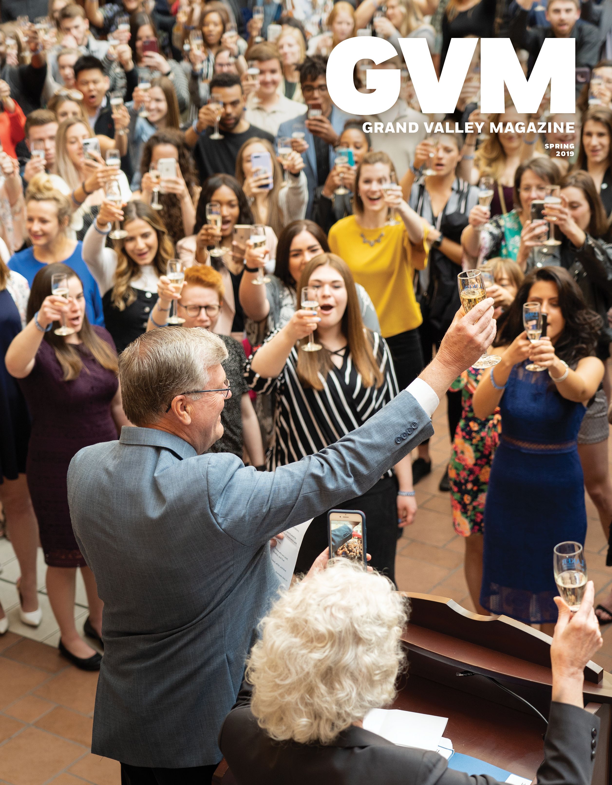 a crowd of people toasting toward a man and women standing on a stage, we are looking at a slightly elevated shot seeing the back of the heads of the man and women on the stage. 