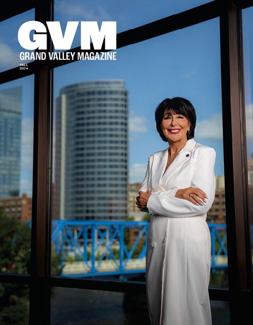 President Mantella dressed in a white coat and GVSU pin is standing with her arms crossed in front of windows, downtown GR (blue bridge and amway tower) are in the background.