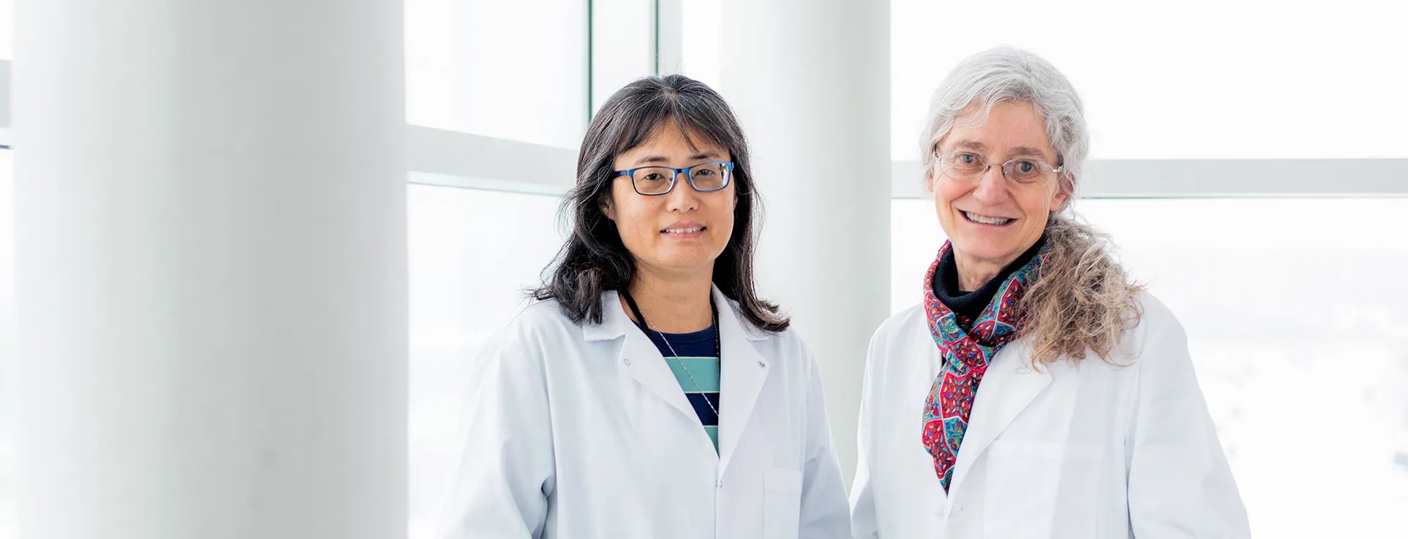 Pei-Lan Tsou, left, and Sheila Blackman stand next to each other in white lab coats, they are standing in front of windows