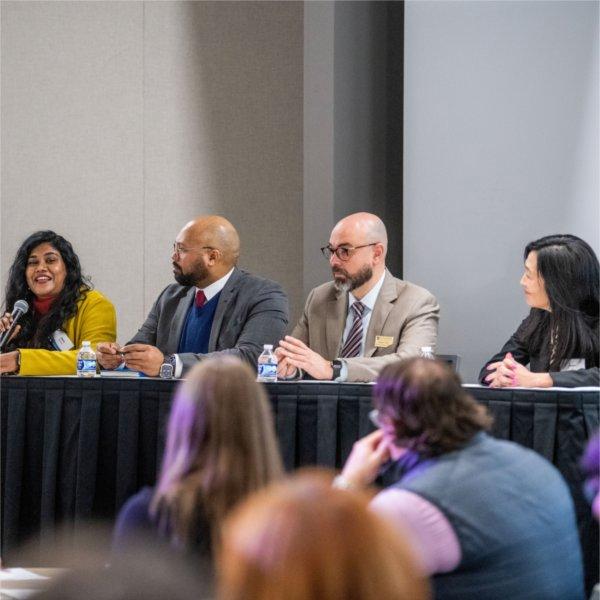 four people are panelists at a table, woman on far left with microphone